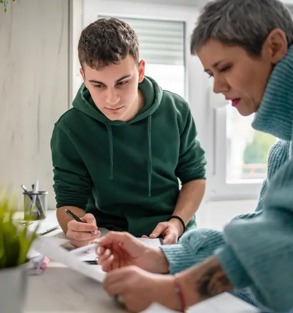 Young man in a green hoodie attentively writing while an older woman with short hair and a blue sweater looks on, at a desk. Bright, focused setting.